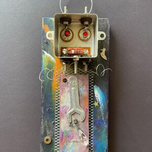 May include: Mixed media artwork on a rectangular wooden base. It features a metal box with red-lensed components, a wrench, and a clear crystal. The background has colourful paint strokes and a wire loop.