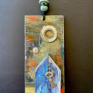 May include: A rectangular, abstract art piece with a blue house shape and a silver key. The artwork features a metal washer, a small gear, and a string of green beads. The background has a textured, colorful paint design.