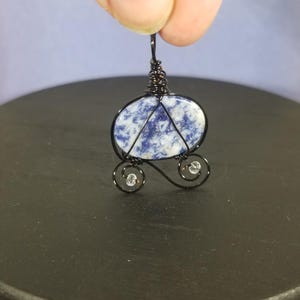 May include: A blue and white stone cabochon is wrapped in black wire to create a pendant in the shape of a carriage. The carriage has two wheels with clear crystal beads.