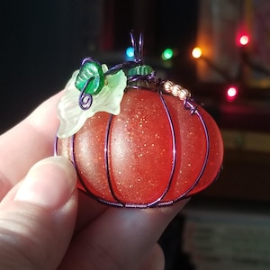 May include: A small, red glass pumpkin with a wire frame and a green leaf accent. The pumpkin is covered in glitter and has a purple wire loop for hanging.