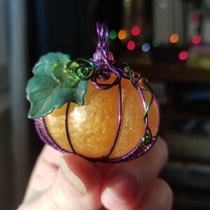 May include: A small, orange pumpkin-shaped ornament with a green leaf and purple wire accents. The ornament is made of resin and has a glittery finish.
