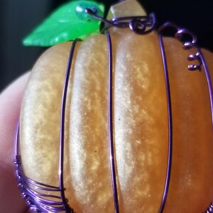 May include: A small orange pumpkin shaped glass ornament with a green leaf and purple wire wrapped around it.