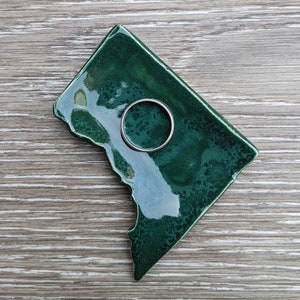 May include: A dark green ceramic ring dish shaped like a state, with a silver ring resting on it. The dish has a glossy finish and is placed on a wooden surface.