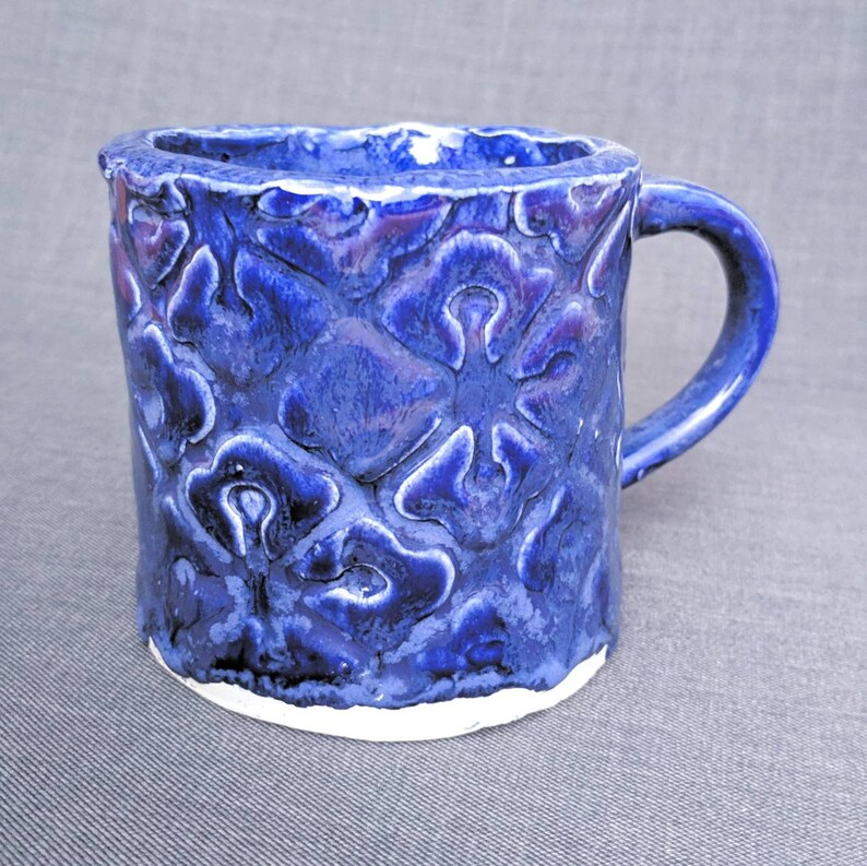 May include: A handmade ceramic mug with a textured, floral pattern in shades of blue. The mug has a white base and a curved handle. The interior and handle are a solid blue, while the exterior features a raised, textured design. The mug is approximately 4 inches tall.