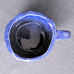 May include: A handmade ceramic mug with a dark blue interior and a textured, lighter blue exterior. The mug has a small handle and a unique, uneven rim. The mug is designed for beverages.
