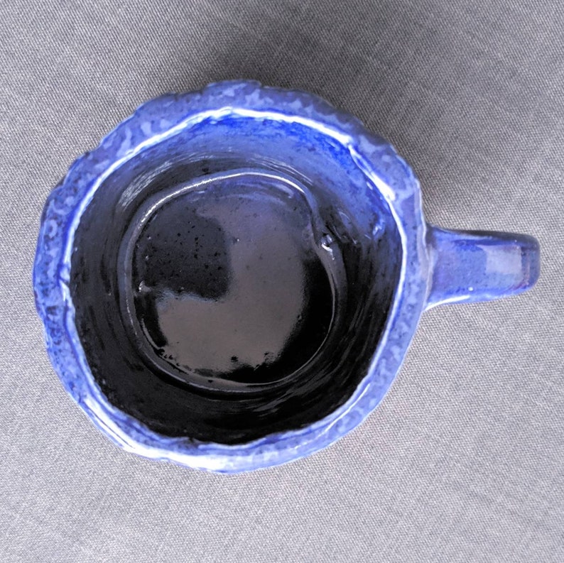 May include: A handmade ceramic mug with a dark blue interior and a textured, lighter blue exterior. The mug has a small handle and a unique, uneven rim. The mug is designed for beverages.
