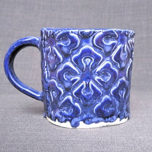 May include: A ceramic mug with a dark blue glaze and a raised floral pattern. The mug has a rounded handle and a white base. The mug is approximately 4 inches tall.