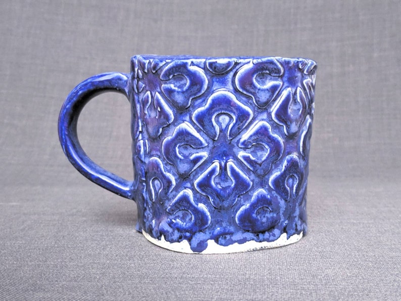 May include: A ceramic mug with a dark blue glaze and a raised floral pattern. The mug has a rounded handle and a white base. The mug is approximately 4 inches tall.