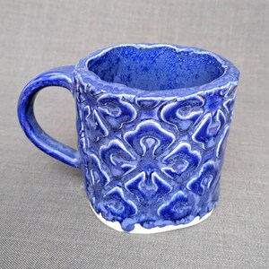May include: A handmade ceramic mug with a deep blue glaze. The mug features a textured, raised floral pattern on the exterior and a contrasting white base. The handle is a matching blue. This unique mug is perfect for coffee or tea.