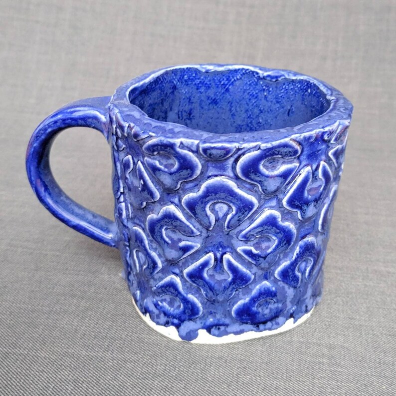 May include: A handmade ceramic mug with a deep blue glaze. The mug features a textured, raised floral pattern on the exterior and a contrasting white base. The handle is a matching blue. This unique mug is perfect for coffee or tea.