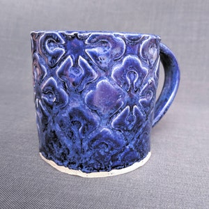 May include: A handmade ceramic mug with a deep blue glaze and a textured floral pattern. The mug has a sturdy handle and a white, unglazed base. The mug is approximately 4 inches tall and 3.5 inches in diameter, perfect for coffee or tea.