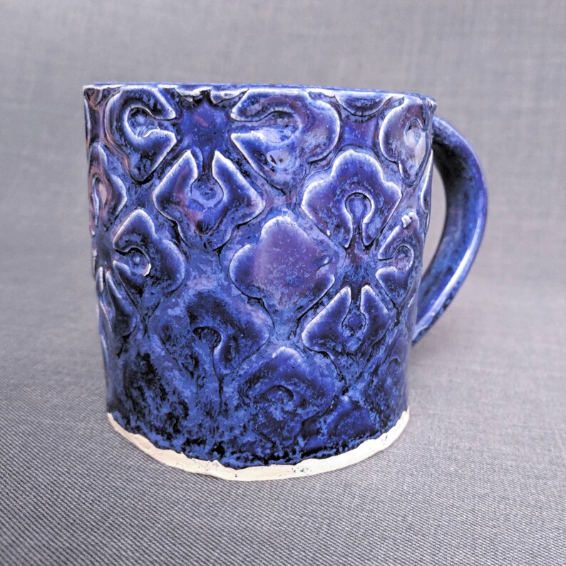May include: A handmade ceramic mug with a deep blue glaze and a textured floral pattern. The mug has a sturdy handle and a white, unglazed base. The mug is approximately 4 inches tall and 3.5 inches in diameter, perfect for coffee or tea.