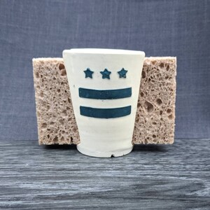 May include: A small, off-white ceramic cup with a dark teal design featuring three stars above two horizontal stripes. The cup is held between two beige, porous sponges, set against a dark gray background.