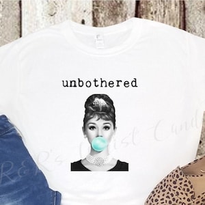 May include: White t-shirt with a black and white image of a woman with a bubble gum in her mouth. The text "unbothered" is printed above the image.