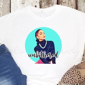 Op de afbeelding: Wit T-shirt met een afbeelding van een vrouw op een blauwe achtergrond met de tekst "unbothered". De vrouw draagt een zwarte turtleneck en een zilveren ketting. Het T-shirt wordt gecombineerd met een blauwe jeans en luipaardprint schoenen.