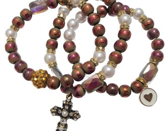 Könnte beinhalten: Drei übereinanderliegende Armbänder mit braunen, schillernden Perlen, weißen Perlen und goldenen Akzenten. Die Armbänder haben einen goldenen Kreuzanhänger und einen Herzanhänger.