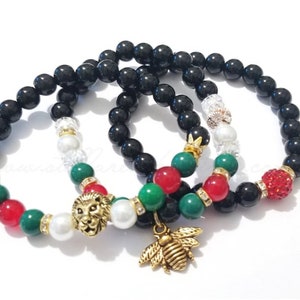 May include: Three beaded bracelets with black, red, green, and white beads. One bracelet features a gold lion charm, another a gold bee charm, and a third a red rhinestone bead. The bracelets are on a white background.