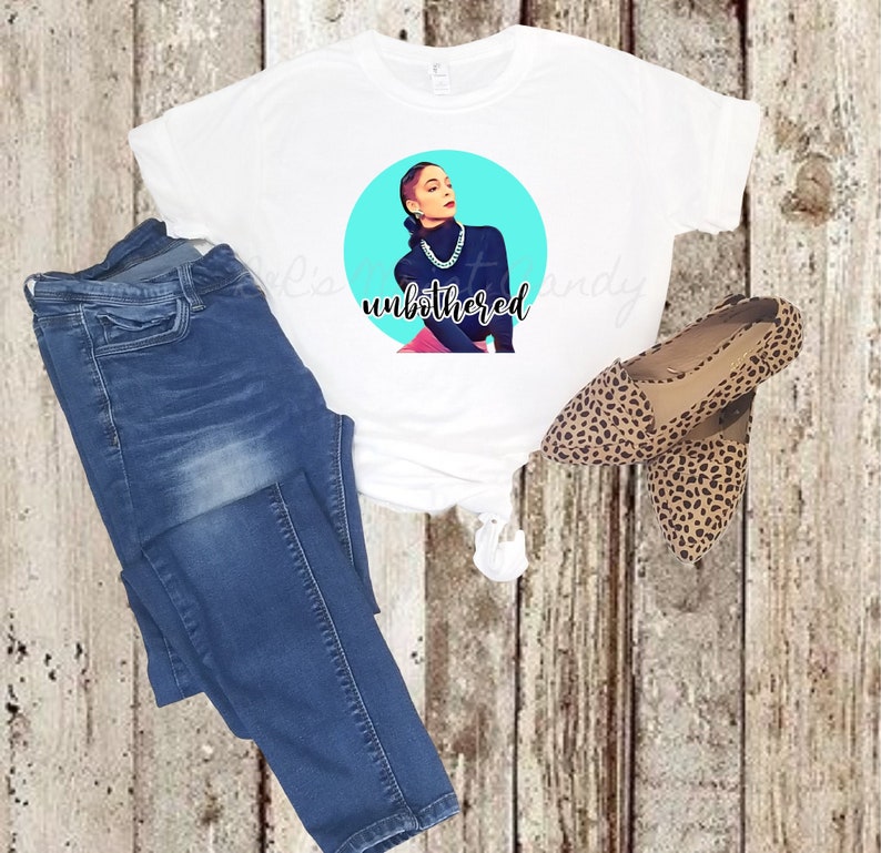 Op de afbeelding: Wit T-shirt met een afbeelding van een vrouw op een blauwe achtergrond met de tekst "unbothered". De vrouw draagt een zwarte turtleneck en een zilveren ketting. Het T-shirt wordt gecombineerd met een blauwe jeans en luipaardprint schoenen.