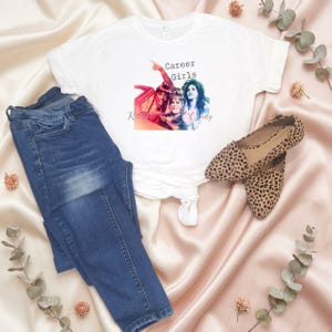 May include: A white t-shirt with the text "Career Girls" and a colorful graphic. The shirt is paired with blue jeans and leopard print flats. The items are arranged on a pink satin surface with decorative elements.