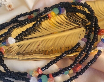 May include: Black beaded waist beads with colorful gemstone accents. The beads are strung together in multiple strands, creating a layered effect. The gemstones include a variety of colors, adding a vibrant touch to the accessory. The beads are displayed on a gold leaf-shaped dish.