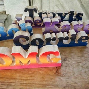 May include: Wooden name signs in various colors, including blue, purple, and orange. The names "Travis", "Karen", "Corrie", and "Brock" are visible, along with the word "OMG". The letters are cut from wood and painted with gradient colors.