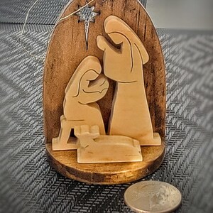 May include: Wooden Christmas ornament depicting the Nativity scene. The ornament features carved figures of Mary, Joseph, and the baby Jesus, with a star above. The ornament is made of light-colored wood and has a dark brown wooden backing. A coin is next to the ornament.