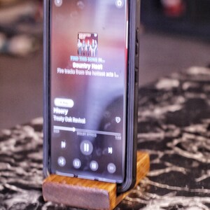 May include: A smartphone in a black case is displayed on a wooden stand. The phone screen shows a music app with the text "FIND THIS SONG IN... Country Heat". The stand is made of light brown wood.