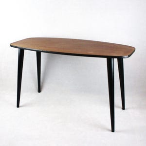 May include: A mid-century modern style coffee table. The tabletop is a light brown color with a black trim. The legs are black and angled, giving the table a sleek, minimalist design. The table is approximately 40 inches long.