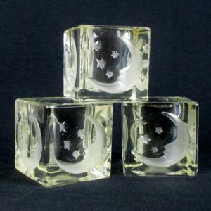 May include: Three clear glass cube candle holders. Each cube features an etched design of a crescent moon with a face and stars. The candle holders are stacked, showcasing the celestial theme.