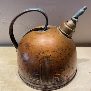 May include: A vintage copper kettle with a blue handle and a bird-shaped whistle. The kettle has a rounded body with a decorative design on the front. The copper has a weathered, aged appearance, adding to its antique charm.