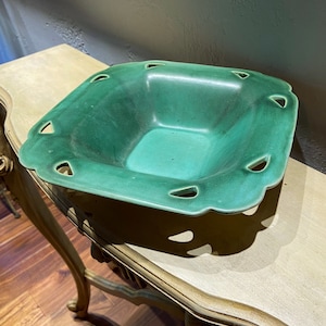 May include: A square green ceramic bowl with cut-out designs around the rim. The bowl is sitting on a light brown wooden surface.