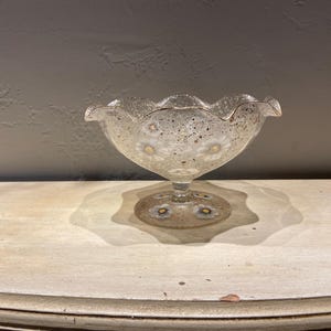 May include: A clear glass footed bowl with a scalloped rim and daisy floral design. The bowl has a speckled pattern and gold trim. The base of the bowl also features daisy designs.