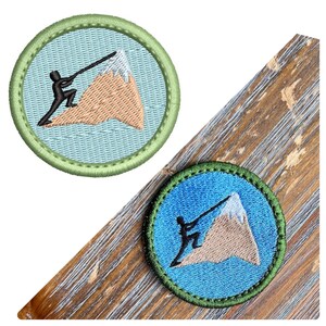 May include: Two embroidered patches with a green border. The patches feature a blue background with a brown mountain and a black figure climbing the mountain. The figure is holding a rope attached to the top of the mountain.