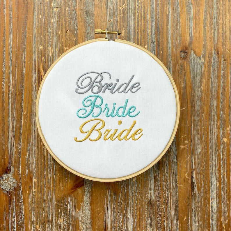 May include: A white embroidery hoop with the word "Bride" embroidered in gray, teal, and gold lettering.