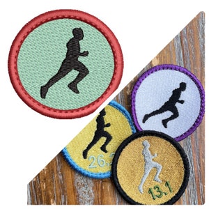 May include: Four embroidered patches featuring a running figure silhouette. The patches are in different colours: red, purple, yellow, and gold. The patches have numbers on them: 26.2, 13.1.