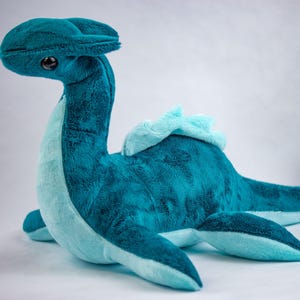 May include: A teal blue plush toy shaped like a sea serpent with a light blue underbelly. The toy has a long neck and a curled tail.