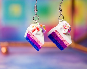 May include: A pair of dangle earrings featuring a slice of cake with blue, pink, and white layers, topped with white frosting and colorful sprinkles.