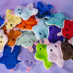 May include: A collection of plush toys in various colors, including blue, yellow, orange, green, and purple. Each toy has a unique design with black button eyes and a pink tongue. The toys are arranged on a purple background.