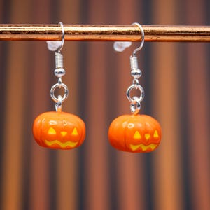 May include: A pair of Halloween-themed earrings. Each earring features a small, orange pumpkin with a carved jack-o'-lantern face. The pumpkins are suspended from silver-colored ear wires. The background is a blurred pattern of orange and brown.