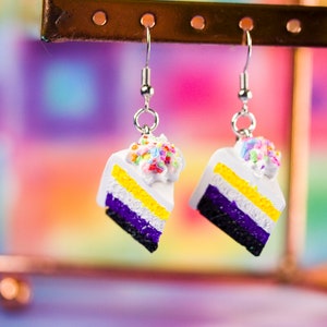 May include: A pair of dangle earrings shaped like cake slices. Each slice features layers of white, yellow, and purple, topped with white frosting and colorful sprinkles. The earrings hang from silver-colored hooks.