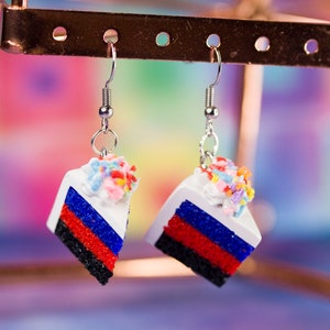 May include: A pair of dangle earrings featuring a miniature cake slice with white frosting and colorful sprinkles. The cake layers are red, blue, and black.