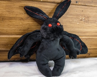 May include: A black plush moth toy with red eyes and wing detailing. The toy features large, bat-like wings and long, pointed ears. The moth is made of soft, fuzzy material and is standing upright.