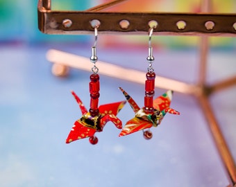 May include: A pair of origami crane earrings. Each earring features a red and patterned paper crane pendant, accented with red beads and silver-tone hooks. The earrings are displayed against a colorful, blurred background.