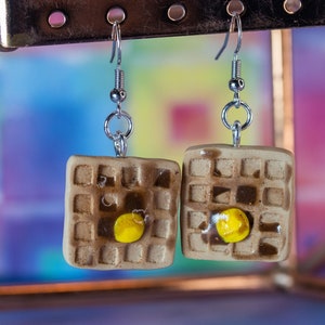 May include: A pair of dangle earrings featuring a miniature waffle with a yellow egg on top. The earrings are made of polymer clay and have silver hooks.
