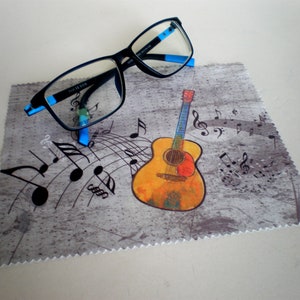 Op de afbeelding: Een grijze microfiber schoonmaakdoek met een kleurrijke gitaar en muzieknoten erop gedrukt. Een paar zwarte brillen met blauwe pootjes ligt op de doek.