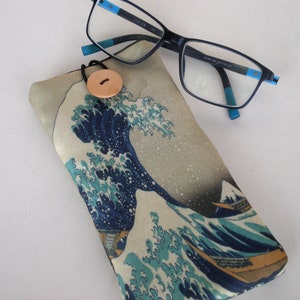 May include: A blue and white fabric glasses case with a wave design and a wooden button closure. The case is lying on a white surface next to a pair of blue-rimmed glasses.