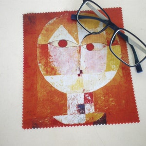 May include: A microfiber cleaning cloth featuring a colorful abstract face design in shades of orange, red, yellow, and white. Black-framed eyeglasses rest on the cloth. The cloth has a decorative, zig-zag edge.