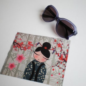 Può includere: Un panno per la pulizia in microfibra grigio e bianco con un design di ispirazione giapponese che presenta una donna dei cartoni animati con i capelli neri e un fiore rosso nei capelli. Il panno ha un motivo floreale rosa e rosso e il testo "YODA" è stampato sul panno.