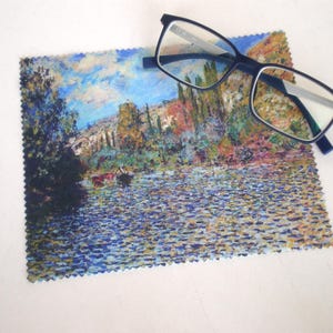 May include: A blue microfiber cleaning cloth with a colourful painting of a river scene. The cloth is folded in half and a pair of blue-framed glasses is resting on top of it.