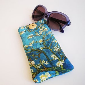 May include: A blue and green floral patterned glasses case with a wooden button closure. The case is lying on a white surface next to a pair of purple sunglasses.